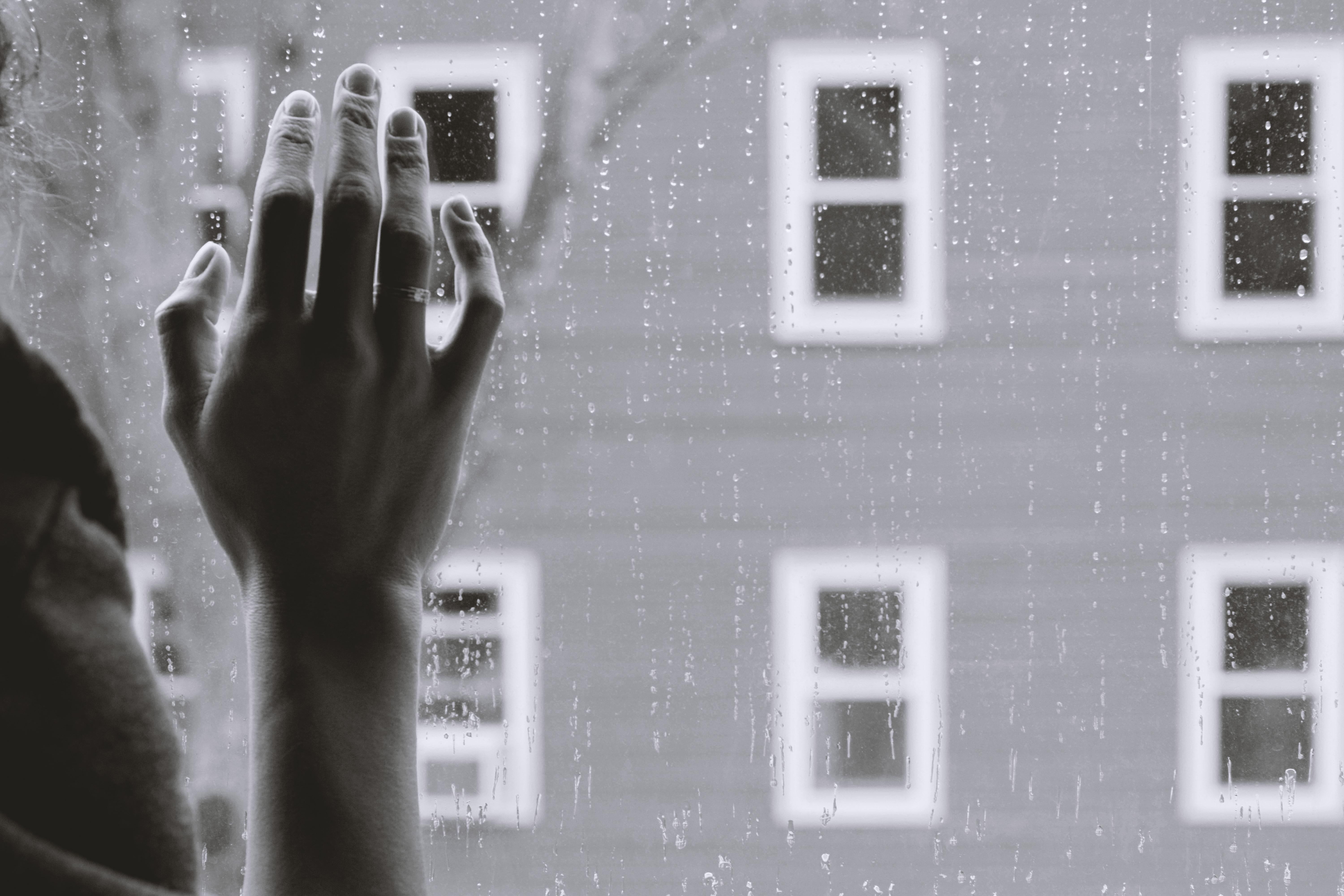 A black and white photo of a woman's hand against a rain-speckled window